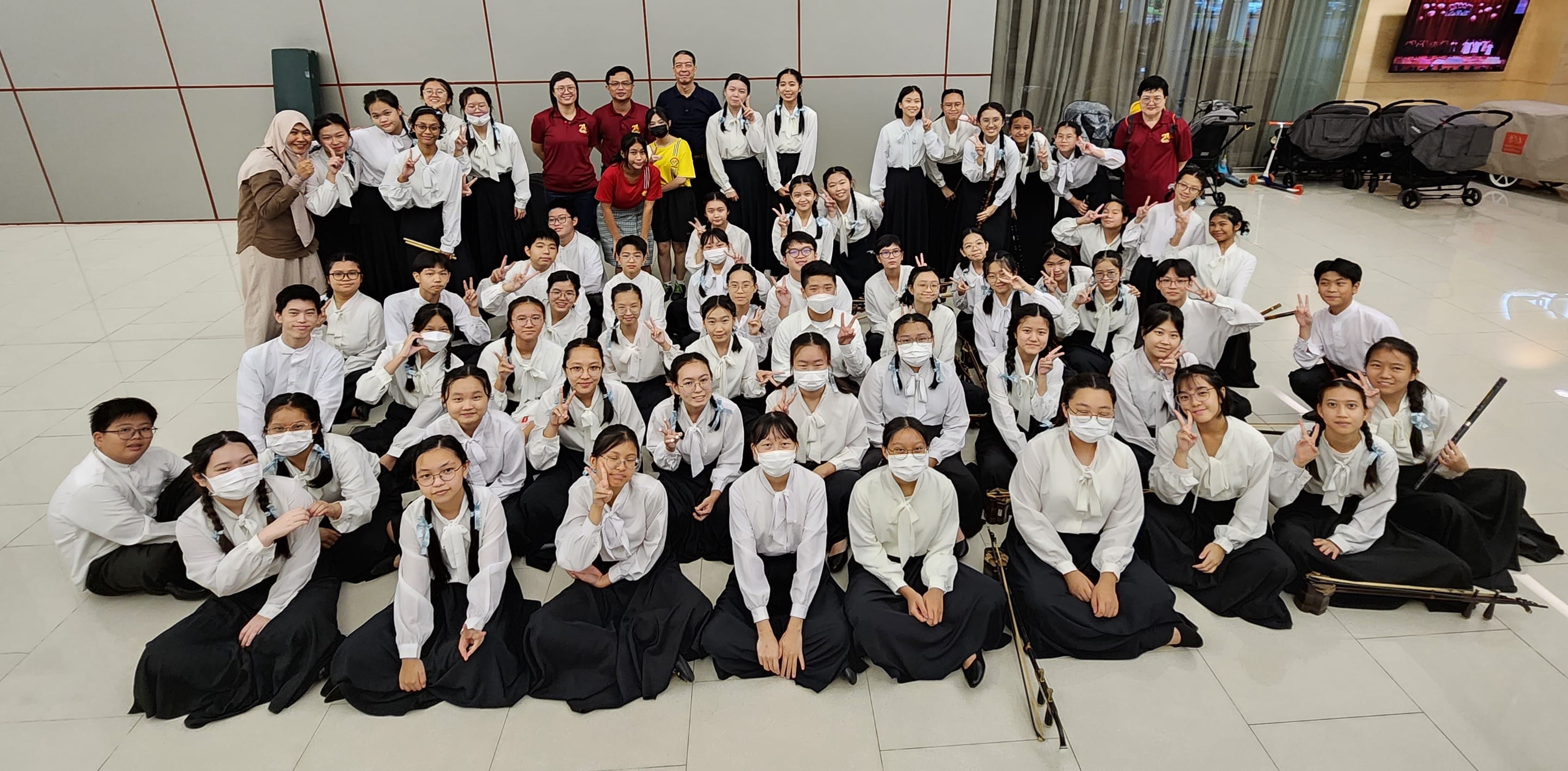 Large group of students in white tops and black skirts posing with peace signs, some wearing face masks, indoors.